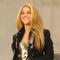 Une femme blanche et blonde souriantequi est Shakira est sur une scène lors de l'inauguration de Barack Obama. Elle est vêtue d'un débardeur doré avec une veste noire, une écharpe noire avec des motifs.