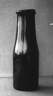 Photo en noir et blanc d’une bouteille, en verre très sombre, dont le goulot est à peine plus étroit que le corps et se termine par un rebord saillant à l’extérieur.
