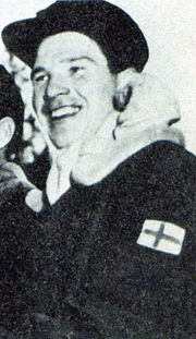 Photographie de Heikki Hasu en 1952. Il sourit et porte une casquette et un manteau avec un drapeau finlandais dessus.