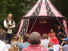 À la droite de la photo se trouve une tente dans laquelle sont assis un homme et une femme. À la gauche, un homme en habit d'époque fait la lecture debout devant des spectateurs assis.