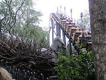 Un manège de montagnes russes s'élève au-dessus des végétations. À gauche, on aperçoit la tête d'aigle grise d'une créature factice.