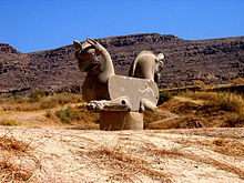 La photo montre une sculpture avec deux têtes de rapaces collées dos à dos, et des pattes antérieures de lion.
