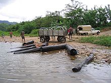 Huit troncs déposés en bord de rivière par des travailleurs riverains, une camionnette et une remorque.