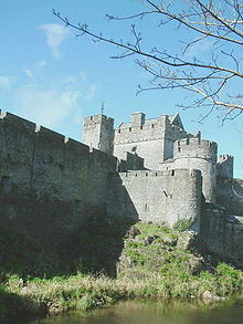 La photographie montre un château-fort surplombant un cours d'eau. Les murs sont surmontés d'un chemin de ronde avec créneaux, et plusieurs tours sont visibles ainsi que le donjon.