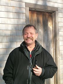 José Bové devant la cabane de Thoreau à Walden Pond. L'écologiste tient une pipe à la main gauche. Debout devant la porte d'entrée de la cabane en bois de Thoreau, il sourit à l'objectif.