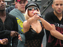 Une femme aux cheveux blonds courts, portant un chapeau de cuir noir ainsi qu'un justaucorps de la même couleur, pointant du doigt un objet alors que plusieurs hommes dansent derrière elle.