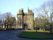 La photographie montre l'entrée d'un château, avec deux hautes tours crénelées entourant une grande porte en bois.