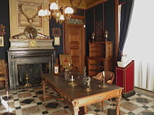 salle au sol et à la cheminée couverts de marbres noir, blanc et rouge, avec une table au centre, des étagères en bois contre un mur de couleur bleu foncé et une tête en marbre posé sur un pupitre en bois
