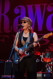 Photographie de Debbie Davis sur scène, debout devant un micro, jouant de la guitare avec une casquette et des lunettes noires.