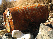 Photo en couleurs d’une canette de boisson rouillée et percée, abandonnée sur un terrain caillouteux.