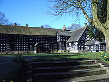 La photographie montre une maison à colombages en forme de L à deux étages. Au premier plan, un escalier en pierre mène à une pelouse, devant la maison.