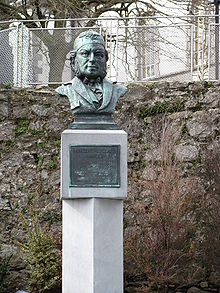 An outdoor bust of a bearded man wearing a large patterned cap and a dicky bow tie. A plaque beneath reads: Vincent Wallace composer. Born Waterford 1812, died Pyrennes 1865. "In happy moments day by day, The sands of life may pass. In swift but tranquil tide away, From times inferring glass."