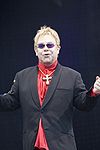 A blond-haired middle-aged man stands with his arms open and fists clenched. He wears a dark suit and red shirt. He also sports a cross pendant around his neck, and wears purple sunglasses.