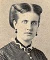 Half-length photographic portrait of a young lady with dark-colored hair swept back and wearing a high-necked, dark Victorian era satin dress with dark buttons