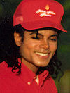 A headshot of a man wearing a red baseball cap and shirt. He has long black hair and is smiling towards the camera.