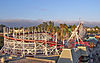 Evening photograph of the white, wooden Santa Cruz Roller Coaster, amusement park structures, and a background of eucalyptus and palm trees.