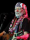 An elderly man holds a guitar while in front of a microphone. He has white facial hair and wears a United States flag bandana.
