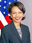 Smiling browned-skinned woman with thickly applied red lipstick wearing a dark blue jacket over a patterned blouse. The United States flag is in the background.