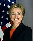 White woman wearing a dark jacket over an orange blouse. The United States flag is in the background.