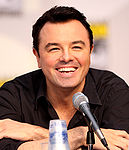A man with black hair and a black shirt, leaning forward, smiling into a microphone.