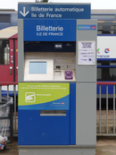 Appareil distributeur automatique de billets de couleurs bleue et grise  portant l'indication « Billetterie automatique Île-de-France », en arrière plan, train de banlieue à deux niveaux.