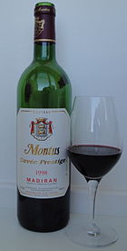 La photographie couleur montre une bouteille de madiran, château Montus prestige millésime 1998. À sa droite, un verre INAO demi-plein montre la couleur pourpre sombre, presque noire du vin.