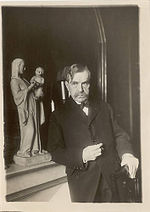 Paul Bourget, moustache, costume sombre, est adossé à une cheminée sur laquelle est exposée la statue d'une vierge à l'enfant. La main gauche du romancier est appuyée sur le dossier d'une chaise. Sa main droite semble tenir une cigarette.