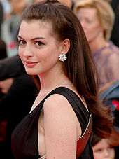A broadly smiling young woman, wearing a white lame strapless gown and small earrings, waves presumably at a crowd. Her is parted on the left side and pulled up and back, off of her bare shoulders. The background is out of focus and unclear.