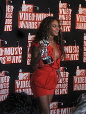 Cette image nous présente une femme brune afro-américaine portant une robe décolleté et courte rouge lors de son passage sur le tapis rouge. Elle a les cheveux bouclés et elle tient de la main gauche un moonman qui est le trophée des MTV Video Music Awards. Le fond est noir avec des logos de la céremonie.