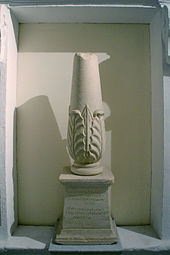Photographie représentant un cippe, petite colonne tronquée en haut, renflée à la base et décorée d’un motif de feuillage.  Elle repose sur un socle trapézoïdal dont la face avant porte les inscriptions - en phénicien au-dessus, en grec en-dessous.