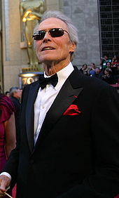 Clint Eastwood porte un costume noir avec une chemise blanche et un nœud papillon noir pour la cérémonie. On peut apercevoir en arrière plan la statue représentant les Oscars qui devance la salle de la cérémonie.