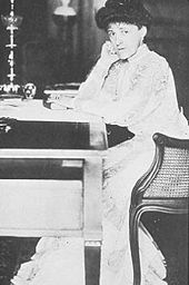 Edith Wharton, assise de trois-quart en train d'écrire, face à une table, chignon et bougeoir sur la table
