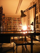Vue de nuit. Des tables de travail avec des lampes de bureau. À l'arrière plan, des rayonnages de livres.