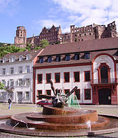 La fontaine est globe sur lequel marchent des êtres humains posée sur un escalier de grès rose en forme d'orbites. Derrière la fontaine, deux maisons dont un bâtiment baroque blanc et rouge surplombées par les ruines du château de Heidelberg.