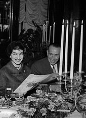 L’image en noir et blanc montre Maria Callas et Pierre Bourgeois souriants, au deuxième plan, derrière une somptueuse table.