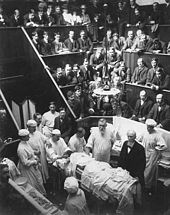 Des étudiants dans un amphithéâtre assistent à une opération, au premier plan le professeur et le personnel médical en blouses blanches.