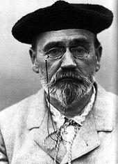 Photographie en noir et blanc d’Émile Zola montrant un homme déjà âgé, à moustache et barbe en pointe poivre et sel, le col noué d’une cordelette, portant des lorgnons et un béret de laine noire.