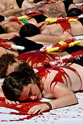 photo couleur montrant un groupe de personnes dénudées étendues sur le sol et simulant la mort, avec du sang et des banderilles sur le dos