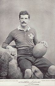 Photo portrait d'Arthur Gould vêtu du maillot gallois. Il est assis et retient de la main un ballon de rugby posé sur sa cuisse gauche.