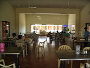 Photographie d’une assez grande salle où sont disposées des groupes de tables avec des chaises en plastique. Des ventilateurs sont suspendus au plafond. Quelques étudiants sont attablés.
