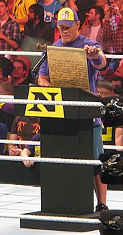 John Cena, nouveau membre du Nexus, se tient debout sur le ring, en face d'un pupitre. Il lit un discours rédigé sur un parchemin.