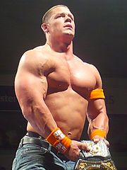 Vue en contre-plongée du catcheur John Cena, septuple champion de la World Wrestling Entertainment. Torse nu, vêtu d'une paire de jeans et de brassards oranges, il tient dans ses mains la ceinture du championnat susnommé, remportée en 2010.