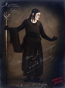Représentation du personnage de Créon dans le costume créé par Coco Chanel : robe noire formée d'une lourde jupe longue et de larges pans de tissu surmontant un justeaucorps à l'encolure échancrée laissant voir la blancheur du cou et terminé par des gants de même couleur.