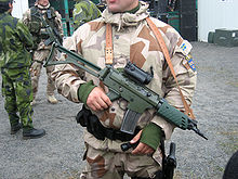 Photographie d'un soldat portant un fusil d'assaut en bandoulière, dont le canon est surmonté d'une lunette à viseur point rouge.