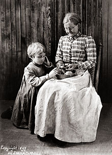 Portrait en noir et blanc d’une femme âgée et d’un enfant posant devant un lambris de bois : la femme, assise, épluche au couteau une pomme de terre au-dessus d’un plat posé sur ses genoux tandis que l’enfant, à genoux sur sa droite, examine un tubercule du récipient. Le maintien de la femme est guindé mais le visage incliné et attentif est légèrement souriant ; elle porte des lunettes rondes et les cheveux gris tirés en chignon, un corsage à carreaux et un ample tablier clair, ligné, qui descend jusqu’au sol. L’enfant aux cheveux courts et blonds, semble très sérieux ; il porte un long tablier à carreaux vichy dont les longues manches sont resserrées aux poignets.