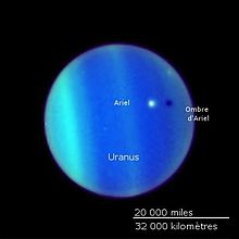 La planète Uranus vue par le télescope Hubble, son atmosphère forme des bandes bleues électrique et vertes. Ariel apparaît comme un point blanc flottant au-dessus et jette un voile sombre au dessous.