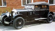 Une Bentley 4½ Litre Sporting Four Seater noire, capote mise, stationnée devant un bâtiment de briques rouges.