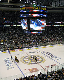 Photo montrant les joueurs des Bruins, les arbitres et les joueurs des Canadiens alignés lors des hymnes sur la patinoire marquée du logo des Bruins.