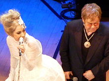 Photographie de Lady Gaga, vêtue d'une robe argentée, chantant sur scène tout en jouant du synthétiseur.