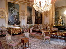 Salle décorée de boiseries blanches et de grandes toiles peintes, d'un tapis, de chaises, d'une commode et d'un bureau